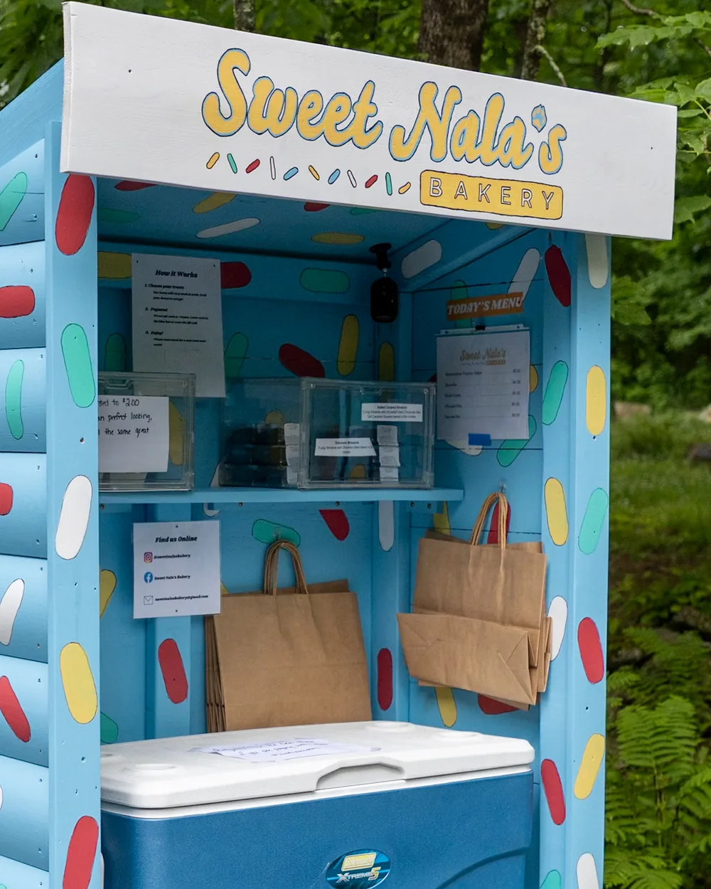 The open bakery cart with kraft paper bags hanging inside and a cooler on the bottom shelf.
