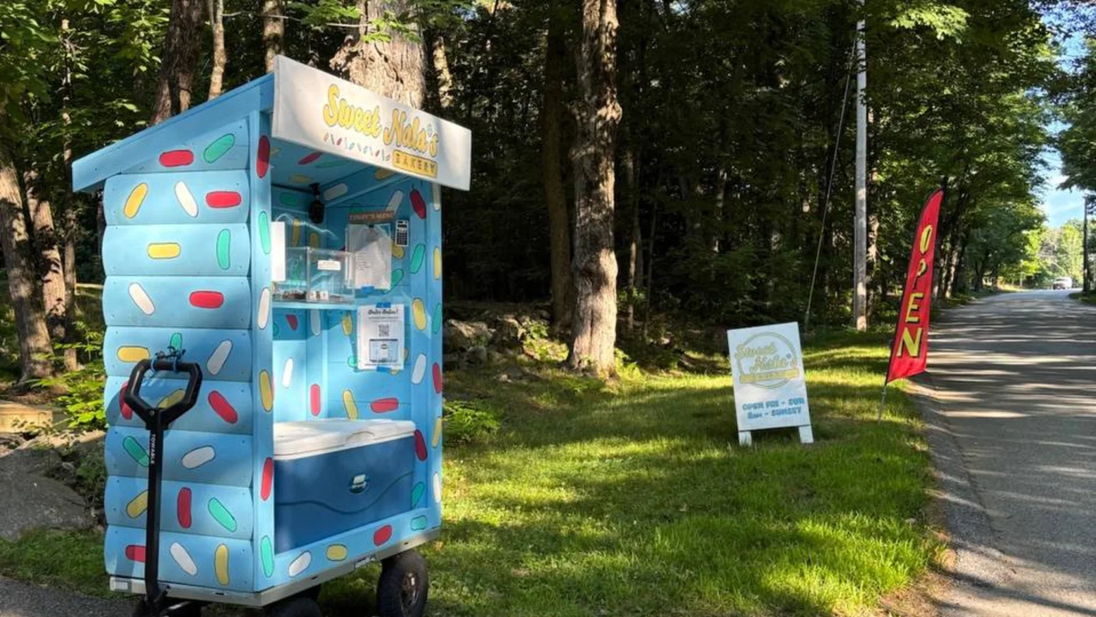The blue bakery cart at the end of the driveway on a summer afternoon, with the white A-frame sign and a red OPEN flag standing beside it at the edge of Ware Corner Road.