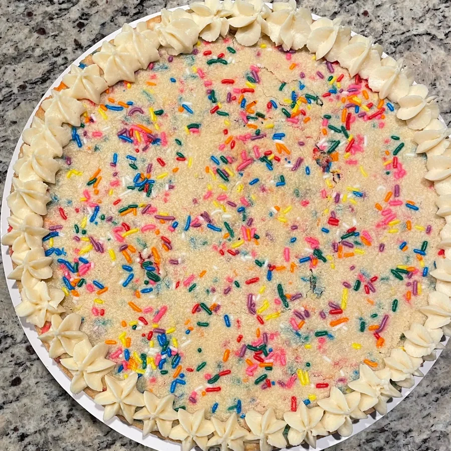 A giant sugar cookie cake covered in rainbow sprinkles with a piped buttercream border.