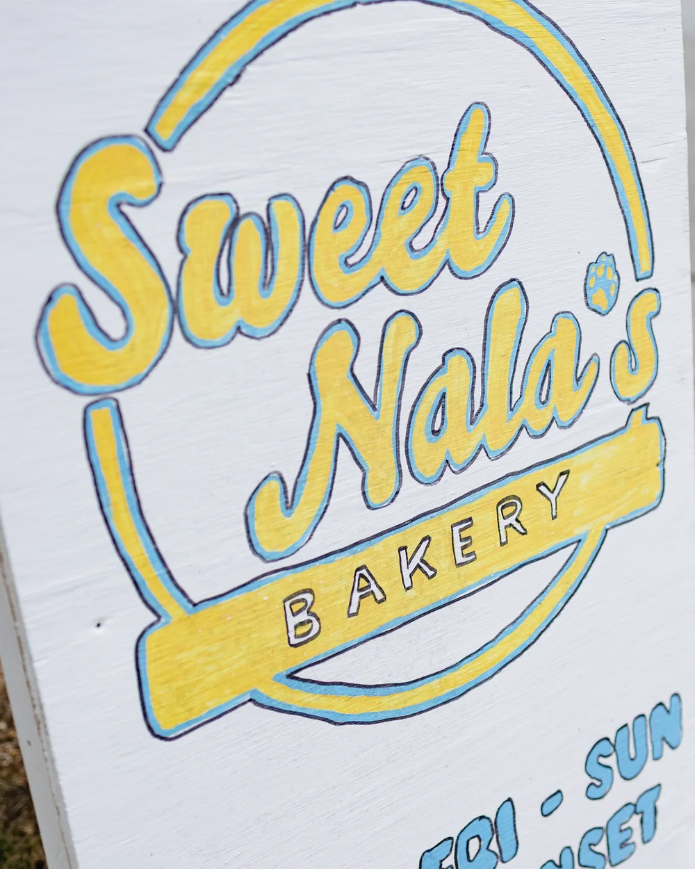 The hand-painted A-frame sign at the roadside: Sweet Nala's Bakery in yellow script inside a circle, with a paw print for the apostrophe, and Open Fri to Sun 8am to sunset lettered underneath.